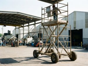 Bouwvakker bedient mobiele rolsteiger op industriële werkplaats met magazijnen en productieapparatuur op achtergrond